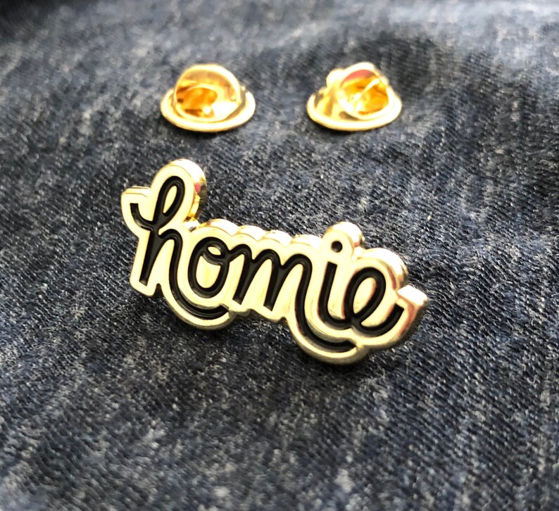 May include: A gold-colored pin with the word "homie" in a cursive font with a black outline. Two gold-colored pin backs are also visible. The pin is set against a dark blue denim background.