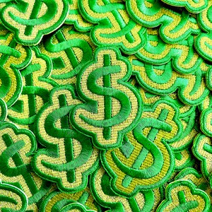 Puede incluir: Primer plano de una pila de parches bordados con el signo del dólar, de color verde y amarillo. Los parches están dispuestos en un patrón aleatorio y todos son del mismo tamaño.