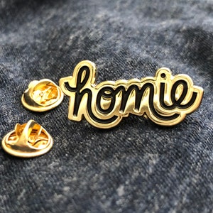 May include: A gold-colored pin with the word "homie" in a black, cursive font. The pin has a shiny, metallic finish and is accompanied by two gold-colored pin backs. The pin is set against a blue denim background.