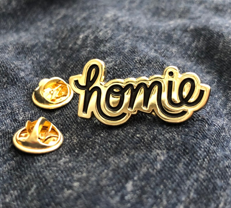 May include: A gold-colored pin with the word "homie" in a black, cursive font. The pin has a shiny, metallic finish and is accompanied by two gold-colored pin backs. The pin is set against a blue denim background.