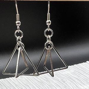 May include: A pair of silver-colored geometric earrings. Each earring features a three-dimensional triangular design suspended from a hook. The earrings are displayed against a black and white textured background.
