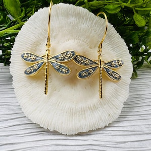 May include: A pair of gold-colored dragonfly earrings. The dragonfly wings are a light blue with gold floral detailing. The earrings are displayed on a white, textured surface with green foliage in the background.