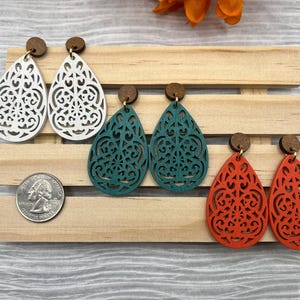 May include: Four pairs of wooden earrings in the shape of a teardrop with a filigree design. The earrings are in white, teal, and orange.