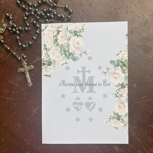 May include: A rosary with a silver crucifix and a card with floral rose accents. The card has the text "A Faithful Love, Blessed by God" and decorative symbols. The rosary beads are dark gray.
