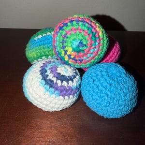 May include: A collection of five colorful, crocheted balls. The balls feature a variety of colors, including blue, green, pink, purple, and white. The balls are arranged on a dark brown surface.