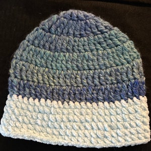 May include: A blue and white crocheted hat with a textured brim. The hat is made with a gradient yarn that transitions from dark blue to light blue.
