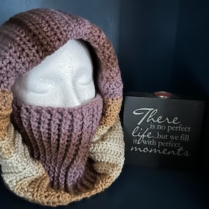 May include: A hand-crocheted hood and cowl set in a gradient of colours, including beige, tan, lavender, and brown. The hood is designed to cover the head and neck, with a built-in face covering. The set is displayed on a mannequin head.