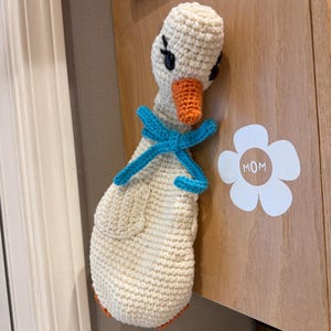 May include: A handmade crochet goose with a cream-colored body, orange beak, and blue bow. The goose has black eyes and is attached to a wooden surface. A white flower sticker with the word "MOM" is also visible.