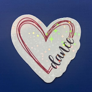 May include: A white heart-shaped sticker with a red outline and the word "dance" written in cursive inside. The sticker has a glittery finish with iridescent stars.