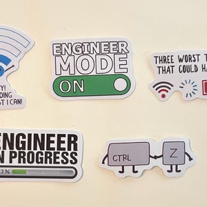 Peut inclure: Cinq autocollants en vinyle sur le th&egrave;me de l'ing&eacute;nierie. L'un montre un symbole Wi-Fi avec le texte "HEY! I'M DOING THE BEST I CAN!", un autre "ENGINEER MODE ON", un troisi&egrave;me "THREE WORST THINGS THAT COULD HAPPEN" avec des ic&ocirc;nes, "ENGINEER IN PROGRESS" avec une barre de chargement &agrave; 23%, et une paire de touches "CTRL Z".