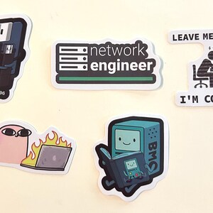 Peut inclure: Cinq autocollants en vinyle sur le th&egrave;me de la technologie : une disquette avec une cl&eacute; USB marqu&eacute;e 1971 et 1996, un logo network engineer, une personne &agrave; un ordinateur avec le texte LEAVE ME ALONE! I'M CODING!, un personnage rose et un ordinateur en feu.