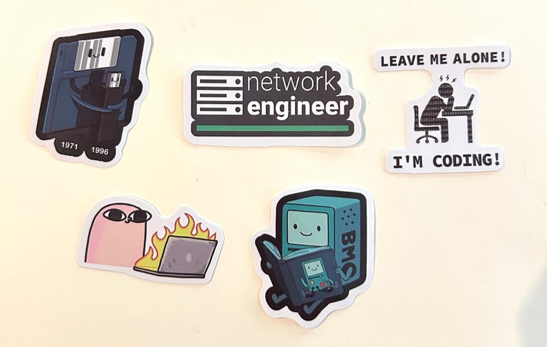 Peut inclure: Cinq autocollants en vinyle sur le th&egrave;me de la technologie : une disquette avec une cl&eacute; USB marqu&eacute;e 1971 et 1996, un logo network engineer, une personne &agrave; un ordinateur avec le texte LEAVE ME ALONE! I'M CODING!, un personnage rose et un ordinateur en feu.
