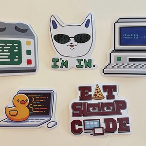 Peut inclure: Cinq autocollants en vinyle sur le th&egrave;me de la technologie : un serveur gris, un chat blanc portant des lunettes &agrave; code binaire avec le texte I'M IN, un ordinateur r&eacute;tro affichant hello world, un canard en plastique jaune sur un ordinateur portable.