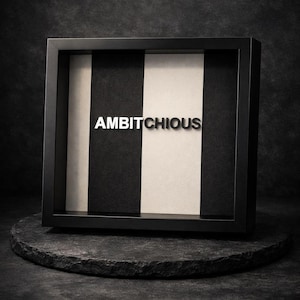 Könnte beinhalten: Ein schwarz gerahmtes Kunstwerk mit dem Wort "AMBITCHIOUS" in silbernen Buchstaben. Der Hintergrund zeigt abwechselnd schwarze und weiße vertikale Streifen. Das Kunstwerk wird auf einem dunklen, strukturierten, kreisförmigen Stein ausgestellt.