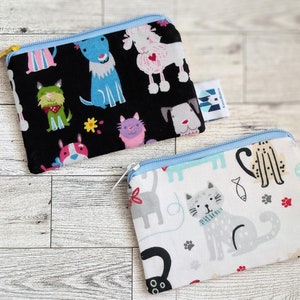 May include: Two small zippered pouches with a black background and a white background. The black pouch has a variety of colorful dogs printed on it. The white pouch has a variety of colorful cats printed on it. Both pouches have a blue zipper and a small tag with the text "Handmade with Love".