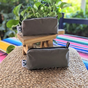 May include: Two gray rectangular zippered pouches with black zippers and small leather pulls. The pouches are displayed on a small wooden stool and a woven mat. Each pouch has a small white tag with a blue and green design.