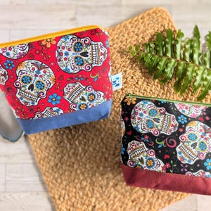 May include: Two colorful zippered pouches with a sugar skull design. The pouch on the left is red with blue trim and the pouch on the right is black with burgundy trim.