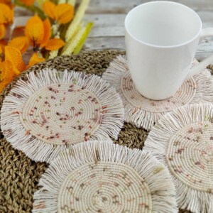 May include: Four crocheted coasters with a fringe border and a speckled pattern in shades of beige, brown, and pink. The coasters are on a woven brown mat with a white mug on top of one of the coasters.