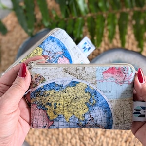 May include: A small zippered pouch with a vintage world map design. The pouch is made of a cream-colored fabric with a blue and yellow map printed on it. The zipper is silver and has a pull tab.