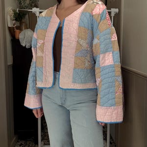 May include: A patchwork jacket in pastel colors, featuring a mix of blue, pink, and tan squares and patterned fabrics. The jacket has a V-neck and is trimmed with blue binding. The quilted design adds texture to this unique outerwear piece.