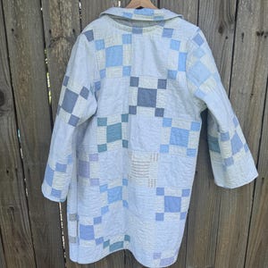 Puede incluir: Un abrigo de manga larga confeccionado con una colcha de retazos que presenta una cuadr&iacute;cula de cuadrados en varios tonos de azul claro, blanco y gris suave. La prenda tiene un cuello y una textura acolchada en toda la tela.