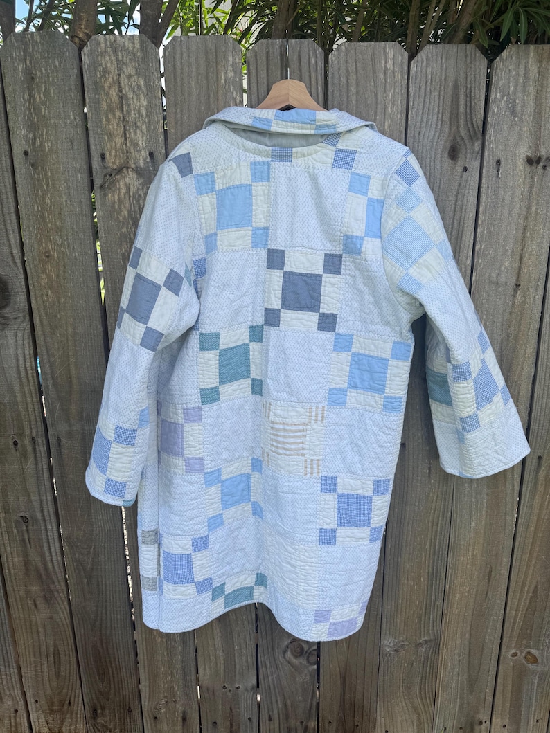 Puede incluir: Un abrigo de manga larga confeccionado con una colcha de retazos que presenta una cuadr&iacute;cula de cuadrados en varios tonos de azul claro, blanco y gris suave. La prenda tiene un cuello y una textura acolchada en toda la tela.