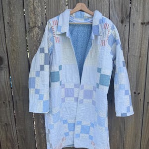 Puede incluir: Una chaqueta de manga larga confeccionada con una colcha de retazos que presenta un patr&oacute;n de cuadr&iacute;cula en tonos blancos, azul claro y morado suave. La prenda tiene cuello y frente abierto, dejando ver un forro interior azul claro con un peque&ntilde;o estampado floral blanco.
