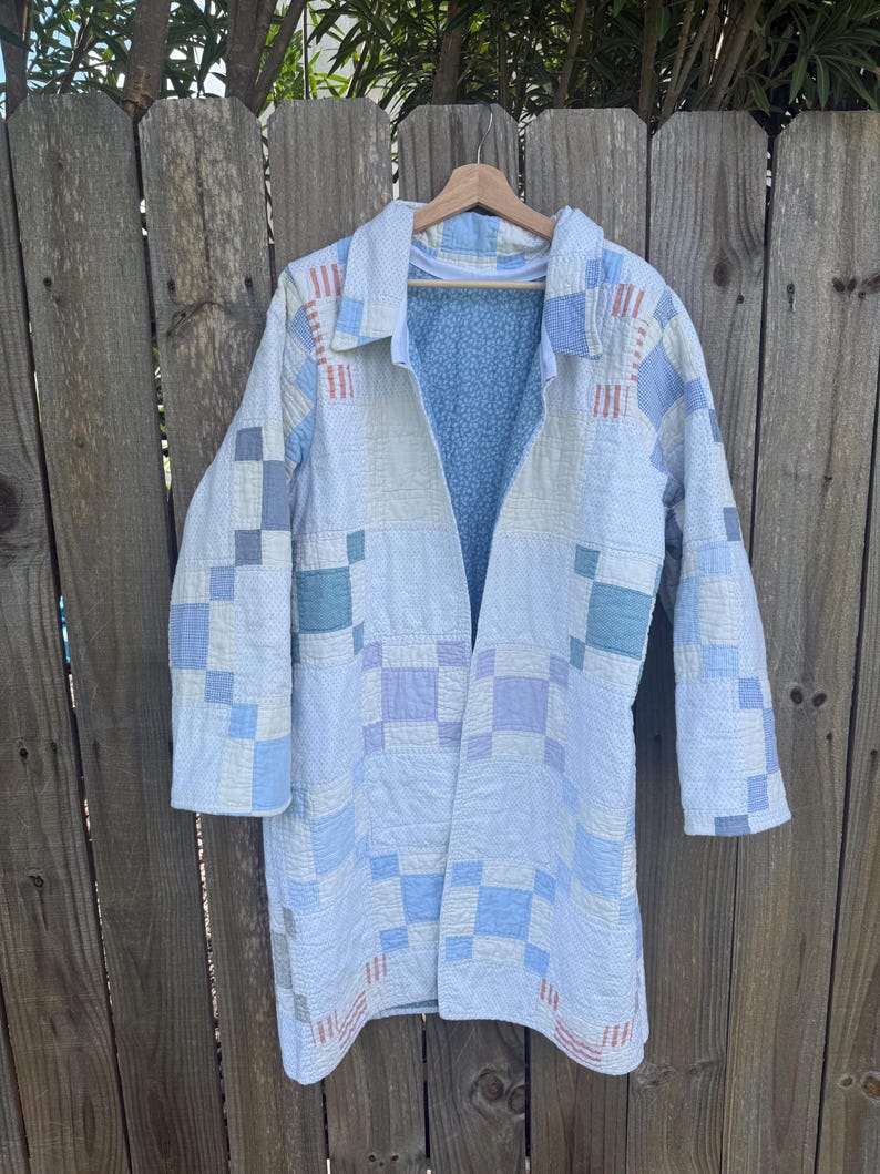 Puede incluir: Una chaqueta de manga larga confeccionada con una colcha de retazos que presenta un patr&oacute;n de cuadr&iacute;cula en tonos blancos, azul claro y morado suave. La prenda tiene cuello y frente abierto, dejando ver un forro interior azul claro con un peque&ntilde;o estampado floral blanco.