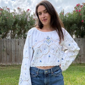 Puede incluir: Un top corto blanco de manga larga con intrincados bordados florales en azul y multicolor, delicados detalles de encaje calado a lo largo de las mangas y el cuerpo, y un dobladillo de encaje festoneado, combinado con vaqueros azules.