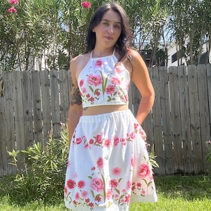 Puede incluir: Un conjunto de dos piezas que consta de un top corto blanco con cuello halter y una falda a juego hasta la rodilla. Ambas prendas presentan un estampado floral vibrante con varias flores rosas y rojas y tallos verdes sobre tela blanca.