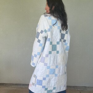 Puede incluir: Una chaqueta de manga larga confeccionada con tela de patchwork acolchada que presenta un patr&oacute;n de bloques repetitivos en tonos blanco, azul claro, gris y verde azulado apagado. La prenda tiene un dobladillo recto y cuello redondo.