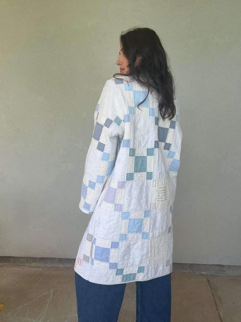 Puede incluir: Una chaqueta de manga larga confeccionada con tela de patchwork acolchada que presenta un patr&oacute;n de bloques repetitivos en tonos blanco, azul claro, gris y verde azulado apagado. La prenda tiene un dobladillo recto y cuello redondo.