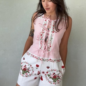 Puede incluir: Un mono sin mangas con un corpiño rosa claro con cierre de cordones en la parte delantera y una sección inferior blanca. La prenda está decorada con un estampado floral y motivos de frutas rojas, incluyendo cerezas y fresas, dispuestas en cestas.