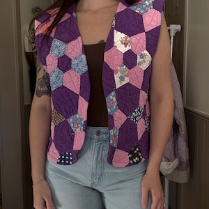 May include: A sleeveless, quilted vest featuring a patchwork design. The vest showcases a pattern of purple, pink, and patterned fabric hexagons. The vest is open in the front and has a V-neckline. The vest is worn over a brown top and light blue jeans.