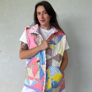 May include: A sleeveless quilted vest constructed from various fabric patches in shades of pink, yellow, blue, and brown. The garment features a front button closure, a collar with a pink and white gingham trim, and two large patch pockets.