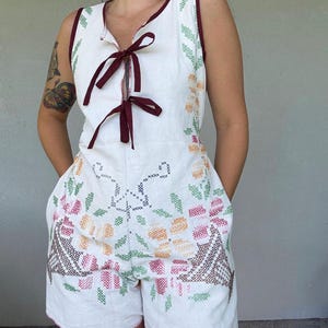 Puede incluir: Un romper blanco sin mangas con un escote profundo y dos lazos de cinta de color rojo oscuro. La prenda está decorada con coloridos patrones de bordado de punto de cruz en tonos verde, naranja, rosa y marrón, e incluye bolsillos laterales.