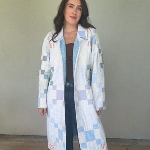 May include: A long, open-front coat constructed from quilted fabric featuring a patchwork pattern of white, light blue, and lavender squares. The garment includes a collar, long sleeves, and a light blue floral-patterned lining visible at the front.