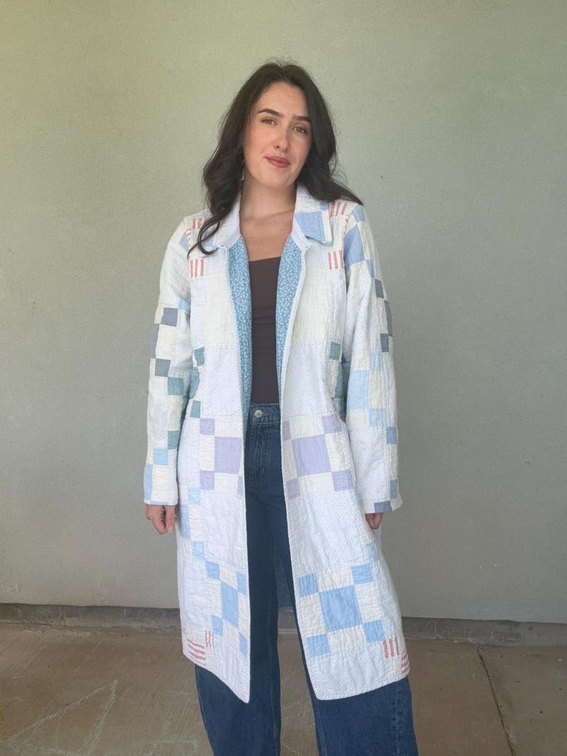 Puede incluir: Un abrigo largo abierto confeccionado en tela acolchada con un patr&oacute;n de patchwork de cuadrados blancos, azul claro y lavanda. La prenda incluye cuello, mangas largas y un forro con estampado floral azul claro visible en la parte delantera.