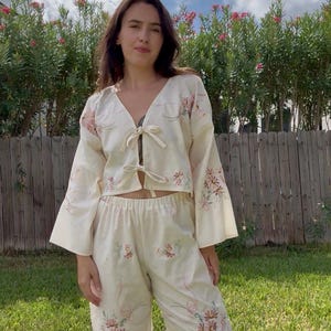 Puede incluir: Un conjunto de dos piezas de color crema que consta de una blusa corta de manga larga con lazos frontales y pantalones a juego de pierna ancha. La prenda presenta un delicado bordado floral en tonos rosa y verde por toda la tela.