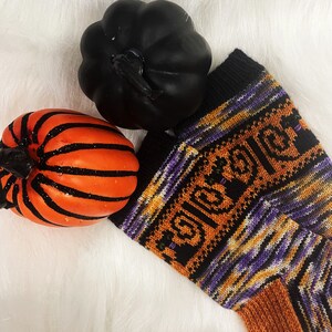 May include: A pair of orange, black, and purple striped knitted socks with the word "Halloween" woven into the design. The socks are lying on a white fluffy surface with two small pumpkins.