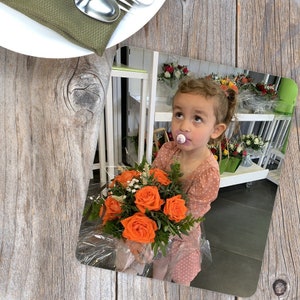 May include: A child holding a bouquet of bright orange roses, wrapped in cellophane with green foliage. The child wears a pink polka-dot dress and looks upwards. The background shows a flower shop display and a wooden table. The bouquet is a lovely gift.