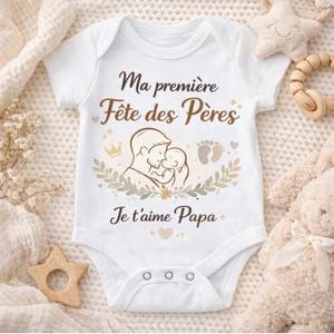 Peut inclure: Body de bébé blanc avec l'inscription "Ma première Fête des Pères" et "Je t'aime Papa" en marron. Le body présente un dessin d'un père tenant un bébé, avec des empreintes de pieds et une couronne de laurier.