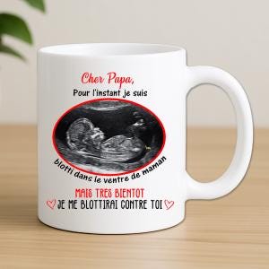 Peut inclure: Un mug en céramique blanc avec une image d'échographie fœtale encadrée d'un ovale rouge. Le texte indique : Cher Papa, Pour l'instant je suis blotti dans le ventre de maman MAIS TRES BIENTOT JE ME BLOTTIRAI CONTRE TOI, avec deux petits cœurs rouges.