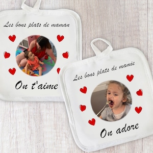 May include: Two white pot holders with red hearts and photos of children. The top pot holder says "Les bons plats de maman" and "On t'aime". The bottom pot holder says "Les bons plats de mamie" and "On adore".