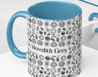 May include: A white ceramic mug with a blue interior and handle. The mug is covered in a black and white pattern of various biological shapes. The text "Dr. Meredith Grey" is printed on the side of the mug.