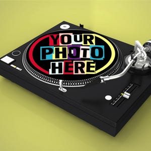 Puede incluir: Plato giratorio negro con un brazo plateado y un gráfico colorido que dice "YOUR PHOTO HERE". El tocadiscos está sobre un fondo amarillo-verdoso. Un diseño clásico para los amantes de la música.
