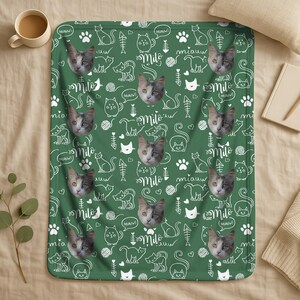 May include: A green fleece blanket featuring a repeating pattern of white line-art cat illustrations, paw prints, fish bones, yarn balls, and the name Milo written in script, interspersed with photographic portraits of a grey and white cat.