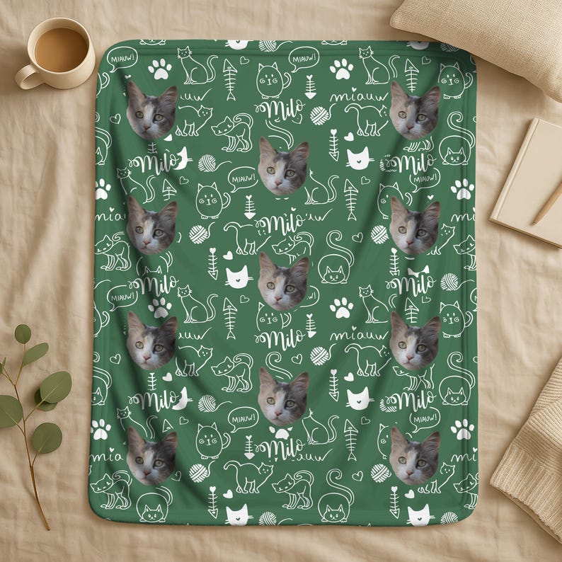May include: A green fleece blanket featuring a repeating pattern of white line-art cat illustrations, paw prints, fish bones, yarn balls, and the name Milo written in script, interspersed with photographic portraits of a grey and white cat.