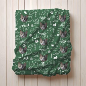 May include: A dark green blanket featuring a repeating pattern of white line-art cat silhouettes, fish bones, yarn balls, paw prints, and the word Milo in script, interspersed with realistic photographic portraits of a calico cat face.