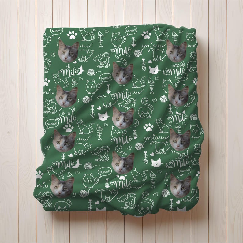 May include: A dark green blanket featuring a repeating pattern of white line-art cat silhouettes, fish bones, yarn balls, paw prints, and the word Milo in script, interspersed with realistic photographic portraits of a calico cat face.